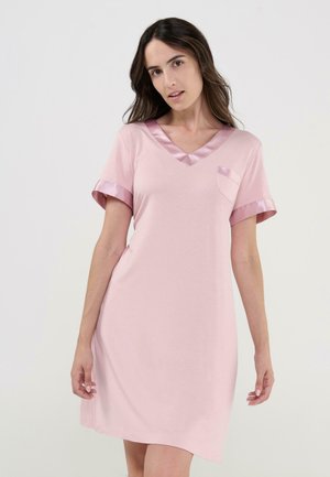 Mujer con camisón rosa de manga corta con ribete de satén en las mangas y escote en V, de pie contra un fondo claro y sencillo.