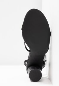 Sandale noire à talon avec une tige texturée, des fines brides autour de la cheville et un talon épais. La semelle est lisse et plate.