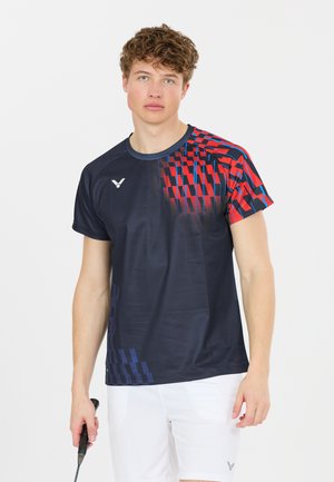 Ung mand iført en sort og rød mønstret sportstrøje og hvide shorts, der holder en badmintonketcher mod en ensfarvet hvid baggrund.