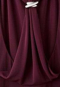 Robe bordeaux avec tissu drapé et un accent métallique torsadé argenté au niveau de l'encolure, présentant une texture lisse et des lignes douces et fluides.