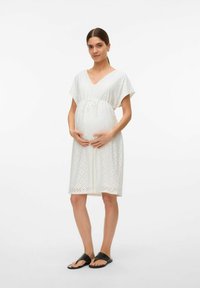 Robe de maternité blanche à manches courtes avec des détails en broderie, un décolleté en V et une taille à cordon de serrage. Portée avec des sandales plates noires.