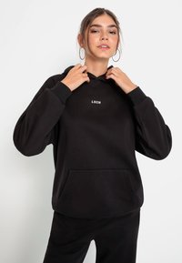 Felpa nera con cappuccio, dotata di tasca frontale e logo bianco "LSCN". Presenta una vestibilità ampia, polsini a costine e una consistenza del tessuto morbida.
