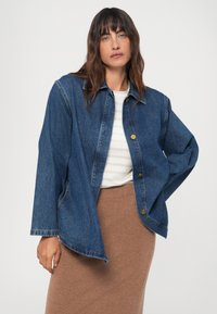 Casaco de ganga com um corte descontraído, cor azul clara, mangas longas e botões dourados. Combinado com uma blusa às riscas e uma saia castanha.