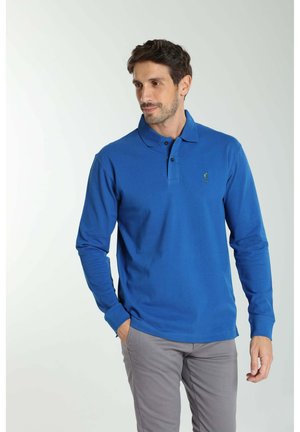 Homme portant un polo bleu à manches longues et un pantalon gris, debout avec une main dans la poche, regardant légèrement sur le côté sur un fond uni.