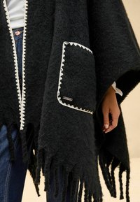 Poncho nero realizzato in tessuto testurizzato, con un bordo bianco a forma di conchiglia, una tasca frontale e bordi frangiati. Indossato sopra un denim.