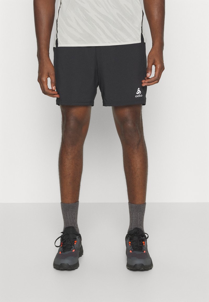 Homme portant un short de sport noir avec un logo blanc, des chaussettes grises et des chaussures de course noires à accents orange, debout devant un fond uni.