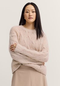 Helles, rosa gestricktes Sweatshirt mit Zopf-Muster, lässiger Passform, überschnittenen Schultern und gerippten Bündchen. Kombiniert mit einem beigen Rock.