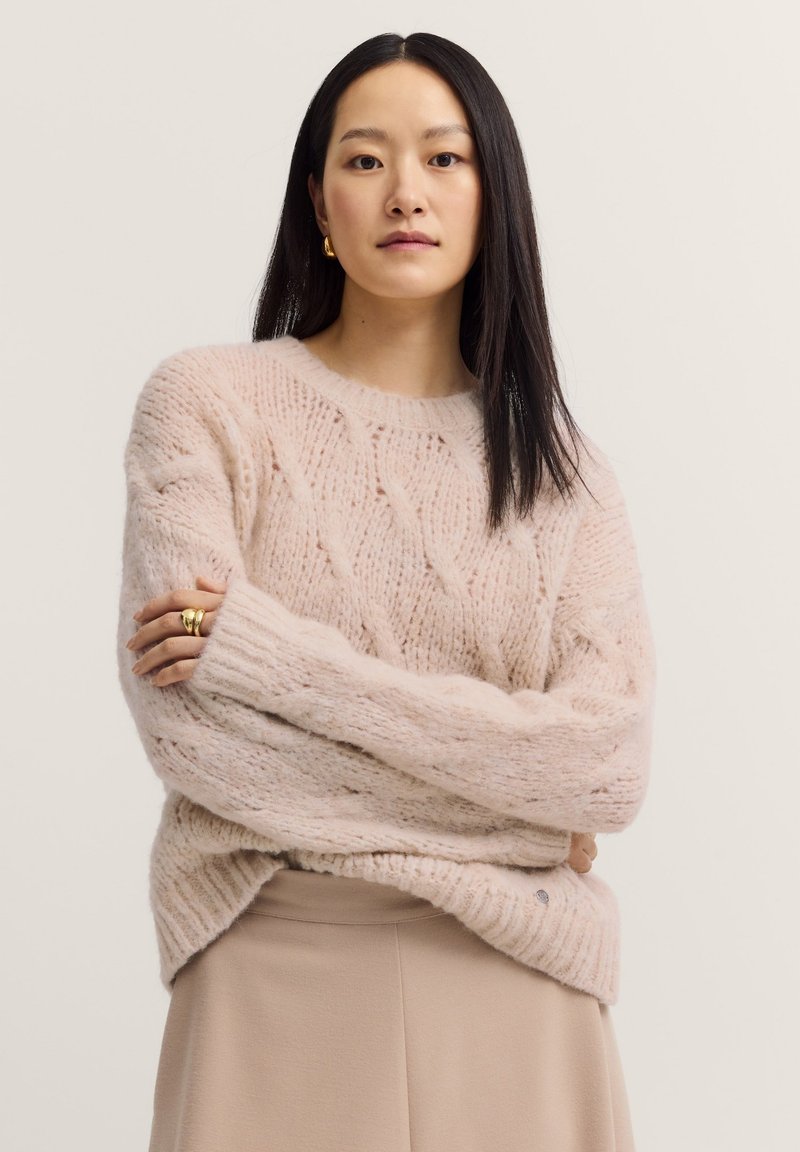 Helles, rosa gestricktes Sweatshirt mit Zopf-Muster, lässiger Passform, überschnittenen Schultern und gerippten Bündchen. Kombiniert mit einem beigen Rock.