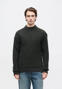 Junger Mann in einem dunkelgrauen Waffelpullover mit einem Schulterreißverschluss und blauen Jeans, steht vor einem schlichten weißen Hintergrund.