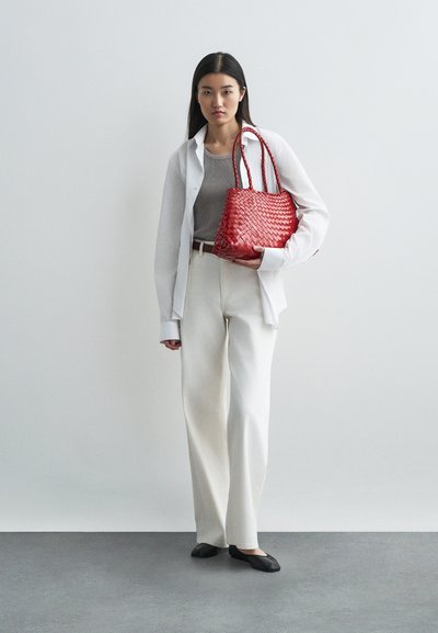 Bolsa de mão tecida em vermelho com duas alças. Modelo veste uma blusa cinza justa, uma camisa branca oversized, calças largas brancas e sapatos pretos rasos. Fundo neutro.