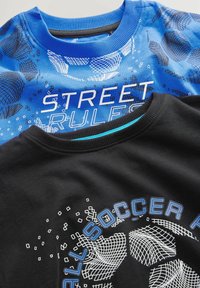 Dos camisetas: una azul con el texto "REGLAS DE LA CALLE" y un patrón de red, la otra negra con un tema de fútbol y detalles azules, ambas de material de algodón.