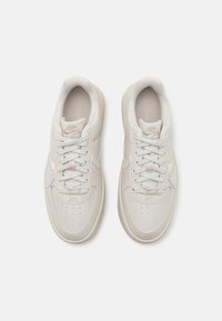 Light beige sneakers with a textured leather upper, flat laces, and a subtle lavender swoosh. Features perforated toe and branding details.