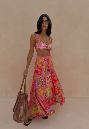 Frau trägt pinkes, geblümtes Croptop und passenden langen Rock, hält eine transparente braune Tragetasche und steht vor einer schlichten beigen Wand.
