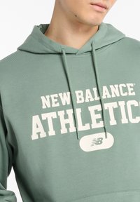 Gray-green hooded sweatshirt with white "NEW BALANCE ATHLETIC" text. Features a front pocket and drawstring hood, made of soft fabric.