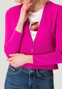 Fuchsiefarbener Cardigan mit Reißverschluss vorne, gerippten Bündchen und Saum; kombiniert mit einem weißen Hemd mit bunten Paillettenakzenten und blauen Denim-Jeans.