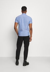 Camicia azzurra a maniche corte con un motivo testurizzato, abbinata a pantaloni neri aderenti e stivaletti neri alla caviglia. Colletto a bottoni visibile.