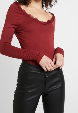 Femme portant un haut bordeaux ajusté à manches longues avec bordure en dentelle et un pantalon en cuir noir avec fermeture éclair à l'avant, mains jointes à la taille.