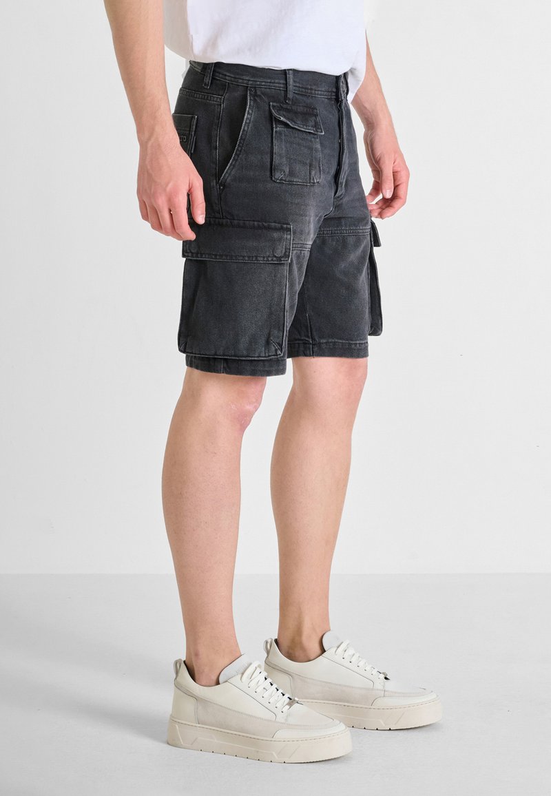 Pantaloni cargo in denim neri con molte tasche, zip frontale e una vestibilità comoda. Abbinati a sneakers bianche con un design testurizzato.