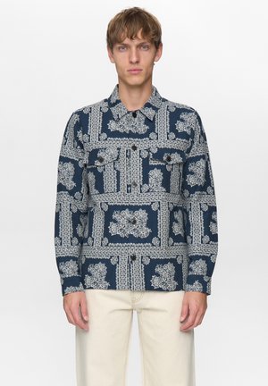 Man draagt een marineblauw overhemd met knopen en witte bloem- en geometrische patronen, gecombineerd met lichtbeige broek, staand tegen een effen achtergrond.