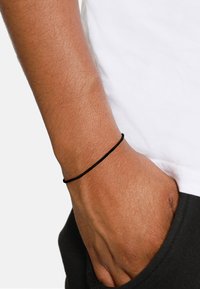 Bracciale elastico nero sul polso, in primo piano. La pelle ha una tonalità marrone chiaro. La mano è appoggiata in una tasca dei pantaloni scuri. Camicia bianca visibile.