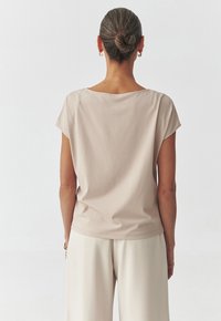 Vaaleanbeige lyhythihainen paita pehmeästä kankaasta, jossa on leveä kaula-aukko ja rento malli. Yhdistettyinä samaan sävyyn beigeihin housuihin. Sileä pinta.