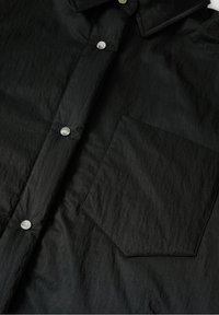 Zwart button-up shirt van textuurstof met een kraag, zilveren drukknoopsluitingen en een borstzak aan de linkerzijde.