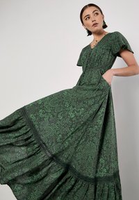 Grön mönstrad maxiklänning med korta ärmar, utsvängd nederkant och fickor. Har intrikata blomsterdesigner och texturerad tyg.