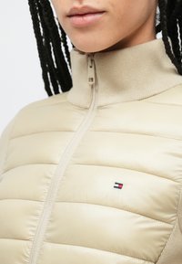 Μπεζ αμάνικο μπουφάν με ψηλό γιακά, με επένδυση από καπιτονέ και εμφανές λογότυπο της μάρκας. Κλείσιμο με φερμουάρ στο μπροστινό μέρος.