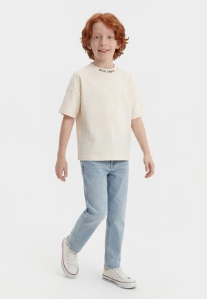 Punapäine laps naeratab, kannab kreemjat Palm Angels t-särki, helesiniseid teksapükse ja valgeid tossusid, kõndides valgel taustal.