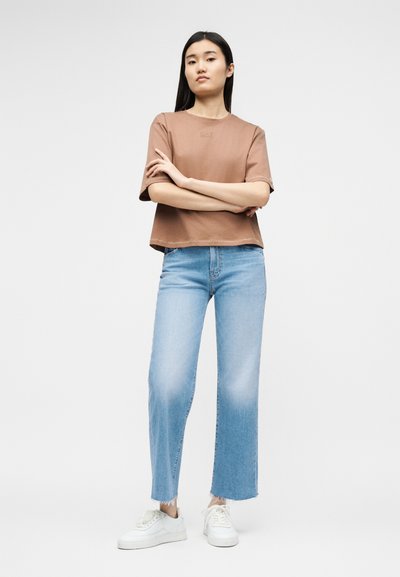 Νεαρή γυναίκα στέκεται με σταυρωμένα τα χέρια, φοράει καφέ κοντομάνικη μπλούζα, ανοιχτόχρωμα τζιν παντελόνια μέχρι τον αστράγαλο και λευκά αθλητικά παπούτσια σε απλό φόντο.