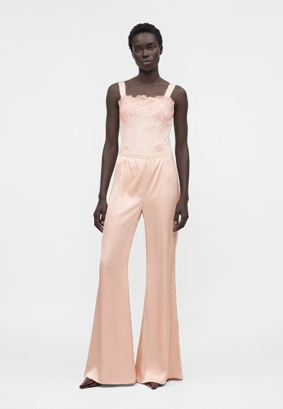 Γυναίκα φοράει αμάνικη παλε ροζ ολόσωμη φόρμα δαντέλα και σατέν, καμπάνα με μυτερά παπούτσια σε απλό ανοιχτό φόντο.