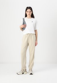 Hvit kortermet t-skjorte med svart logo, beige joggebukser med elastisk midje, grå crossover-veske og lyse sneakers.