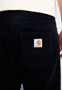 Tamsiai mėlyni kordūrų kelnės su tekstūruota paviršiumi, turinčios baltą pleistrą su logotipu ant užpakalinio kišenės ir oranžinį emblemą.