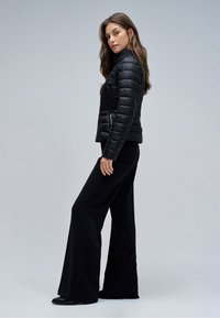 Frau mit langen Haaren, die eine schwarze gesteppte Jacke, schwarze Hosen mit weitem Bein und schwarze Schuhe trägt, seitlich vor einem schlichten grauen Hintergrund stehend.