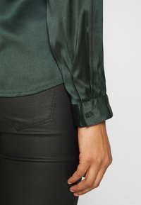 Camicia di raso verde scuro con maniche lunghe e polsini abbottonati. Indossata con pantaloni neri aderenti, che mettono in evidenza la trama liscia e il design sartoriale.