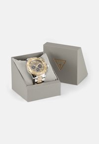 Guld- och silverur i en grå låda, med en texturerad guld bezel, kristallaccenter och svarta urtavlor. Mjuk kudde inuti lådan.
