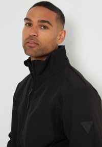 Schwarze Jacke mit hohem Kragen aus weichem, strukturiertem Stoff; verfügt über einen Vorderreißverschluss und ein Logopatch auf der linken Schulter.