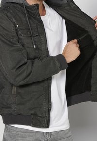 Homme portant une veste vert foncé et une chemise blanche ouvre sa veste pour montrer la doublure en polaire noire et la fermeture éclair de la poche intérieure.