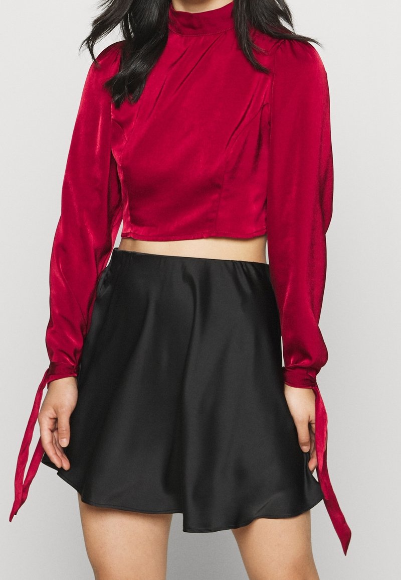 Červený saténový top s krátkými rukávy a mašlemi, v kombinaci s černou saténovou sukní do zvonku. Hladká textura; přiléhavý střih.