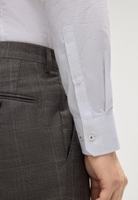 Chemise blanche avec des rayures bleu pâle subtiles, poignets à boutons ; pantalon à carreaux gris avec une texture lisse et une poche latérale.