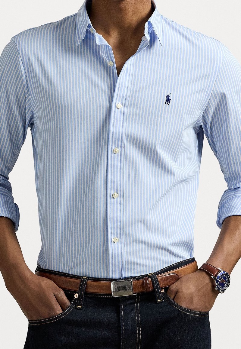 Homme portant une chemise à rayures bleu clair et blanc avec les manches retroussées, une ceinture marron, un jean foncé et une montre-bracelet.