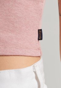 Top court en maille rose clair à côtes avec une étiquette sur le côté. Présente un design ajusté et des coutures plates. Associé à un short en tissu blanc.