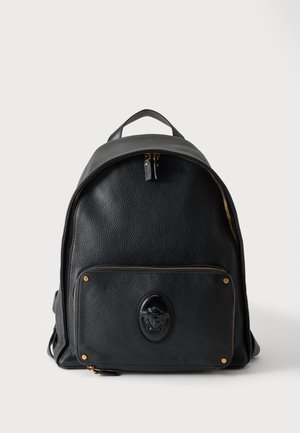 Sac à dos en cuir noir avec une poche avant zippée, présentant un emblème en relief et des détails de fermeture éclair et rivets dorés.