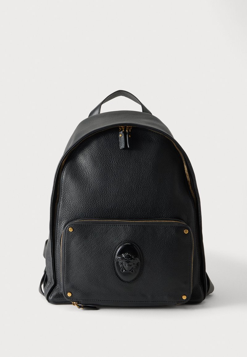 Sac à dos en cuir noir avec une poche avant zippée, présentant un emblème en relief et des détails de fermeture éclair et rivets dorés.