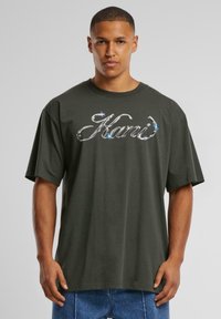 Dark green oversized t-shirt with a round neckline and short sleeves; features a metallic, reflective logo on the front.