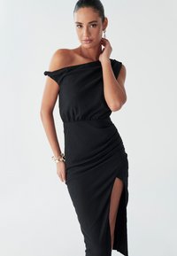 Female model poses confidently in a black off-shoulder dress with a high slit, styled with bracelets and a sleek ponytail against a white backdrop.