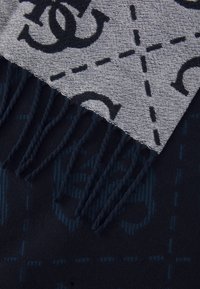 Vilnos šalikas su dviem pusėmis: viena pusė pilka su juodais logotipais, kita tamsiai mėlyna su subtiliu raštu ir pūkuotais kraštais.