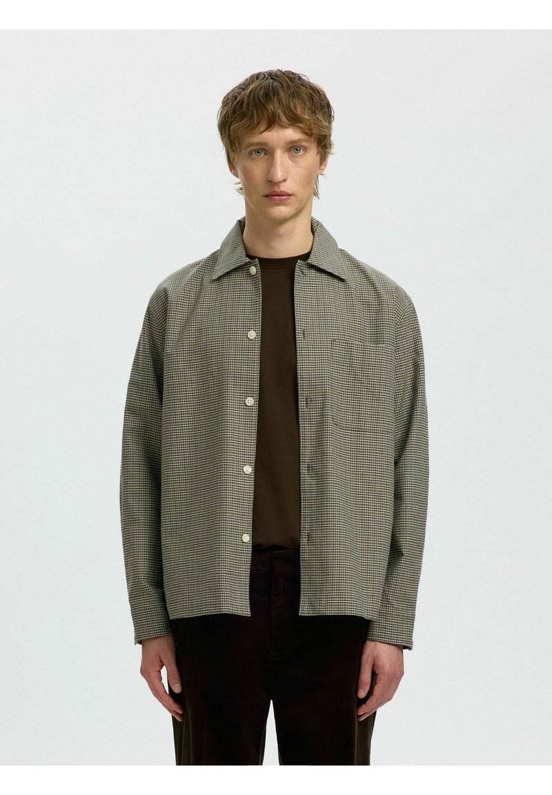 Jeune homme aux cheveux courts et ondulés portant une chemise noire sous une veste grise à carreaux boutonnée avec un pantalon sombre, debout devant un fond uni.