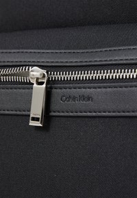 Sac en tissu noir avec fermeture éclair, doté d'un tirette en métal argenté. Le nom de la marque "Calvin Klein" est embossé sur un accent en cuir lisse.