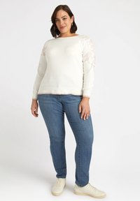 Maglione bianco con ricami floreali sulle maniche, abbinato a jeans in denim blu e sneakers beige. Design semplice, texture morbida.